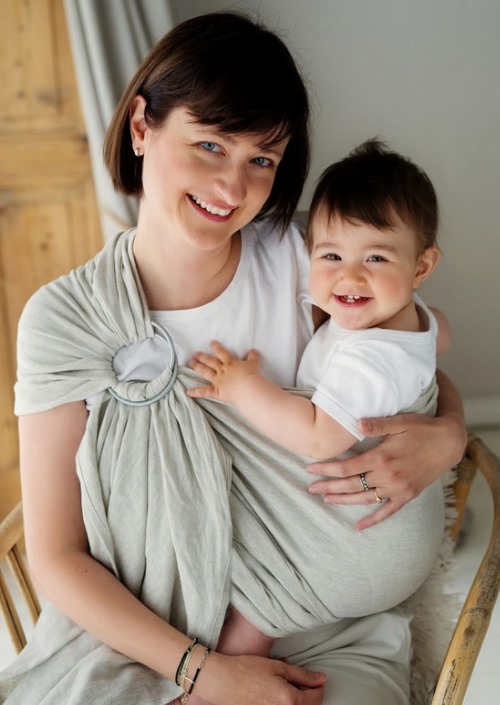 Vrouw draagt een kindje in een lichtgrijze ringsling met metalen ringen op de schouder