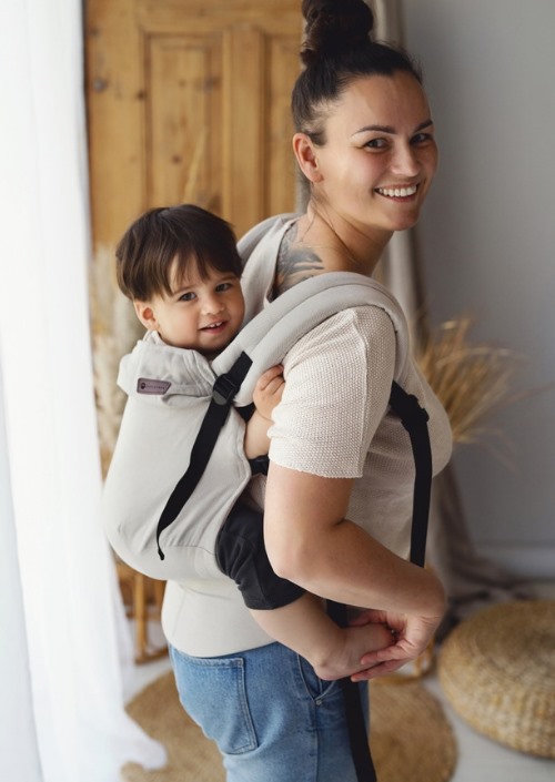 Vrouw draagt een kind op de rug in een beige Little Frog Swift Toddler Carrier met zwarte gespen en banden