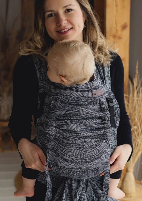 Vrouw draagt baby in grijs-zwarte Little Frog Cross Hybrid Carrier met golvend patroon aan de voorkant