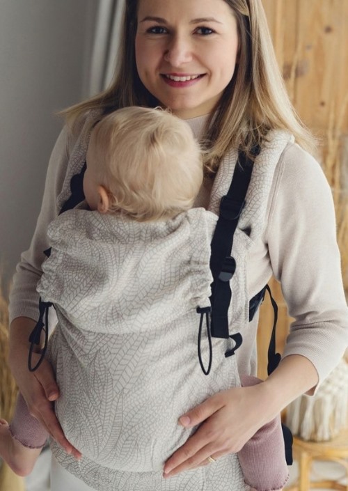 Vrouw draagt een kind op de buik in een lichtgrijze Little Frog Prime Carrier met fijn bladpatroon en zwarte gespen