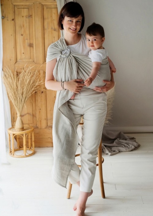 Vrouw draagt baby in lichtgrijze ringsling met ring op de schouder
