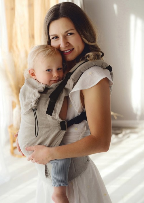 Vrouw draagt baby voorop in beige Little Frog Prime draagzak met zwarte gespen