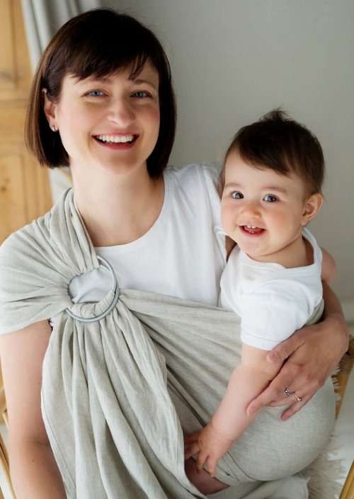 Moeder draagt baby in lichtgrijze ringsling met ringen op de schouder