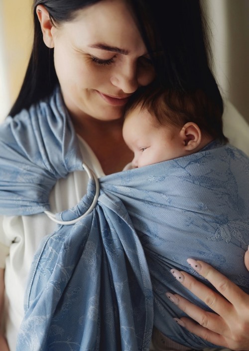 Moeder draagt baby in lichtblauwe Little Frog-ringsling met subtiel bladpatroon en zilverkleurige ringen