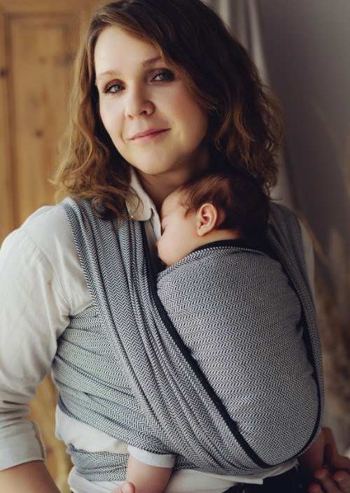 Vrouw draagt een baby in een grijze draagdoek met fijn visgraatpatroon.