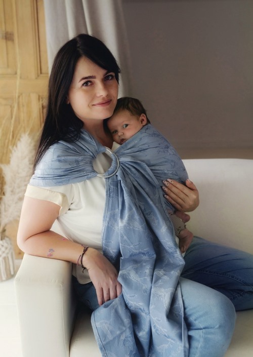 Moeder draagt baby in lichtblauwe Little Frog-ringsling met subtiel bladpatroon en zilverkleurige ringen