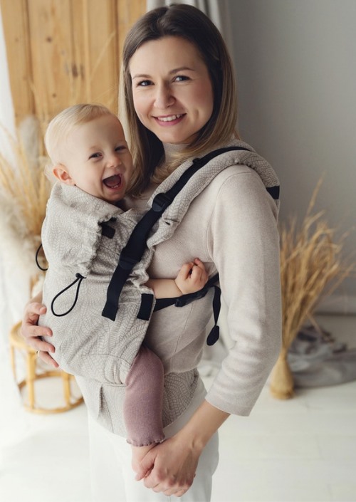 Moeder draagt baby voorop in beige Little Frog Prime Carrier met subtiel bladpatroon en zwarte gespen
