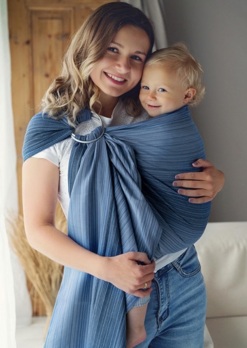 Moeder draagt kind in blauwe Little Frog-ringsling met subtiele strepen en metalen ringen op de schouder